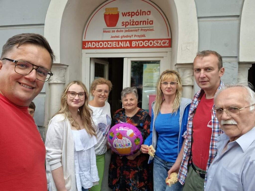 Jadłodzielnia wspólna spiżarnia świętuje Międzynarodowy Dzień Dobroczynności. Wolontariusze na tle jadłodzielni.